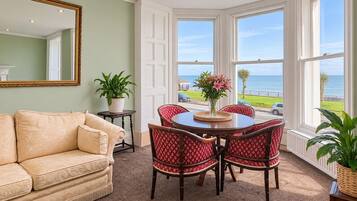A room with a beige sofa, a dining table with red chairs, a large window with a view of the ocean, and a mirror on the wall.