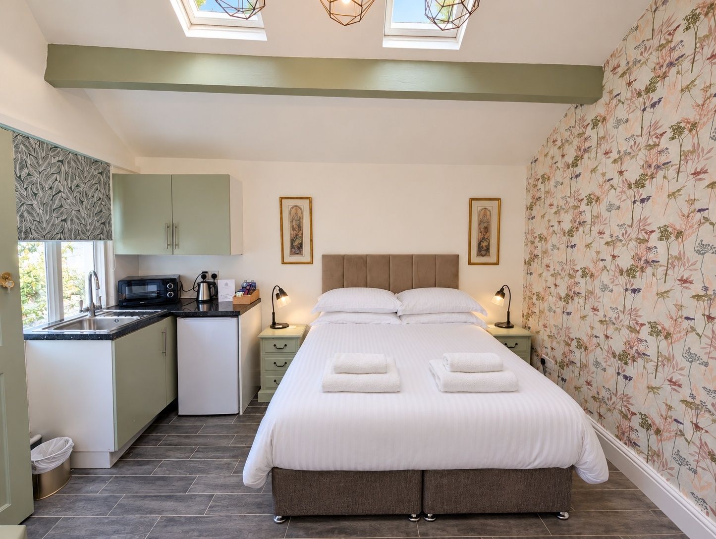 A bedroom with a bed, bedside tables, a nightstand, a window, a small kitchenette, and a bathroom.