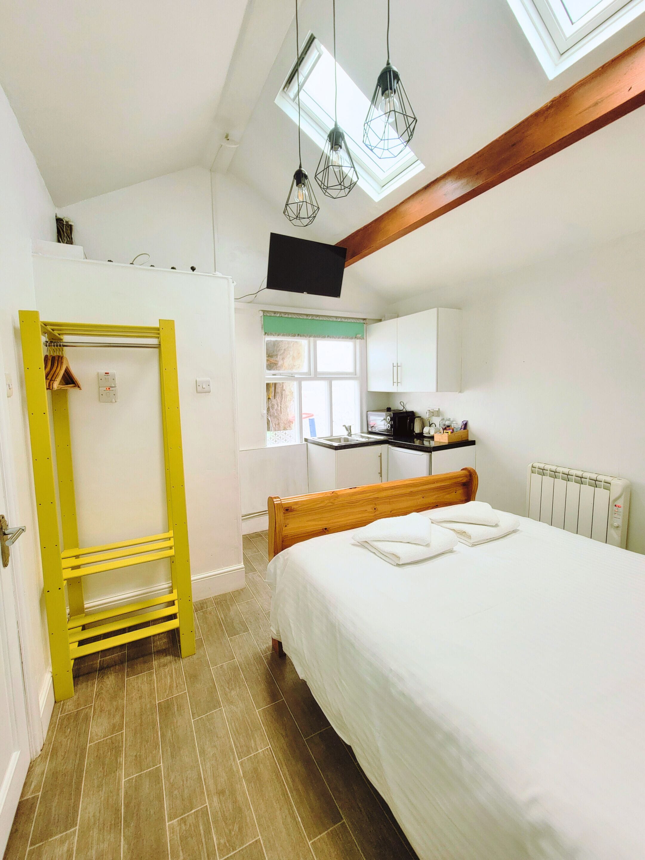 A bedroom with a bed, a yellow shelf unit, a window with a view, and a kitchen area.