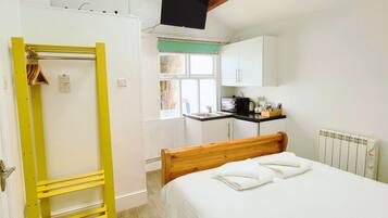 A bedroom with a bed, a yellow shelf unit, a window with a view, and a kitchen area.