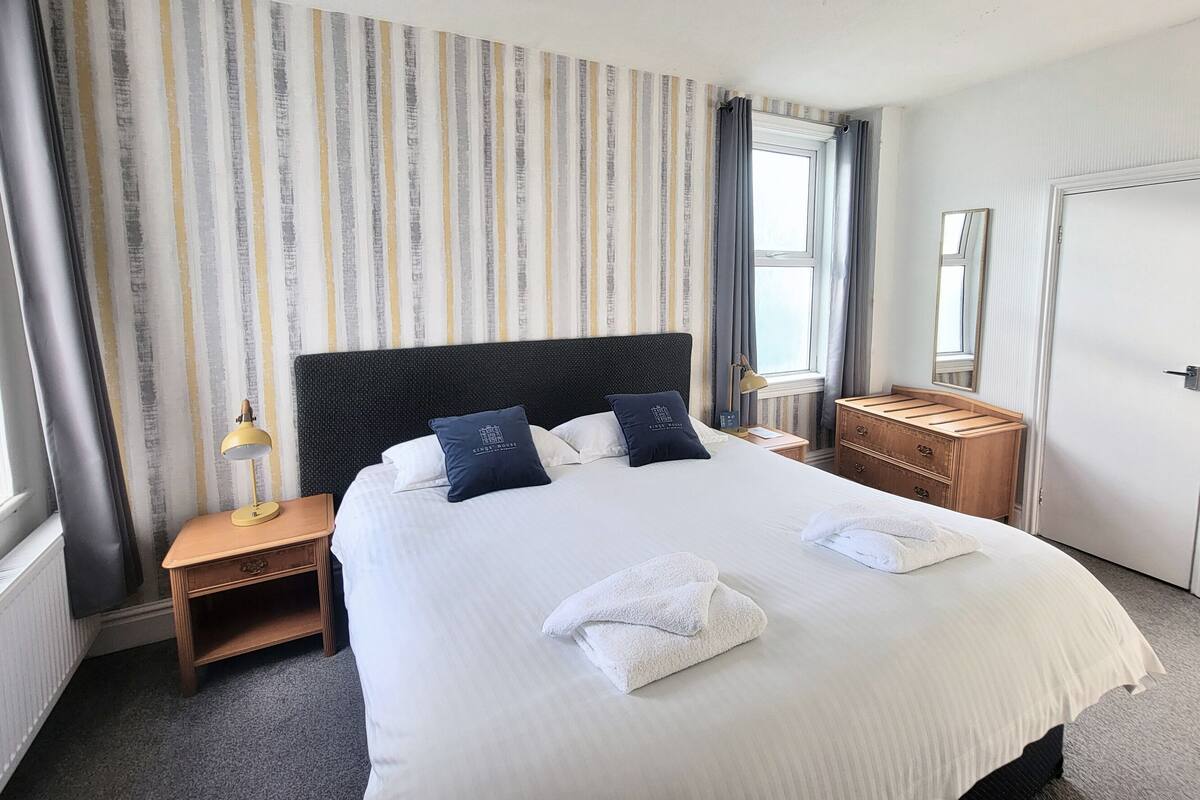 A neatly made bed with two pillows, a bedside table with a lamp, a wooden dresser, and a door.