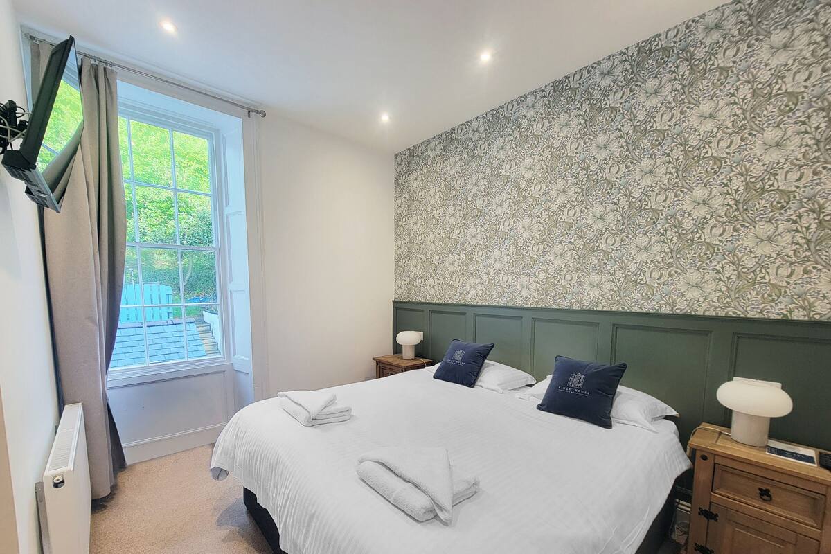 A neatly made bed with pillows, a bedside table with a lamp, and a wall with a floral pattern.