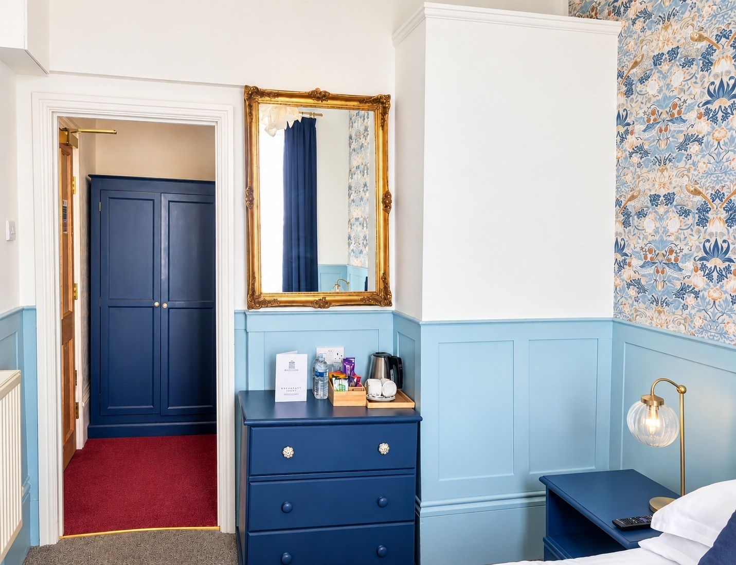 A hotel room with a blue wardrobe, a mirror with a gold frame, a bedside table with a lamp, and a floral-patterned wall.
