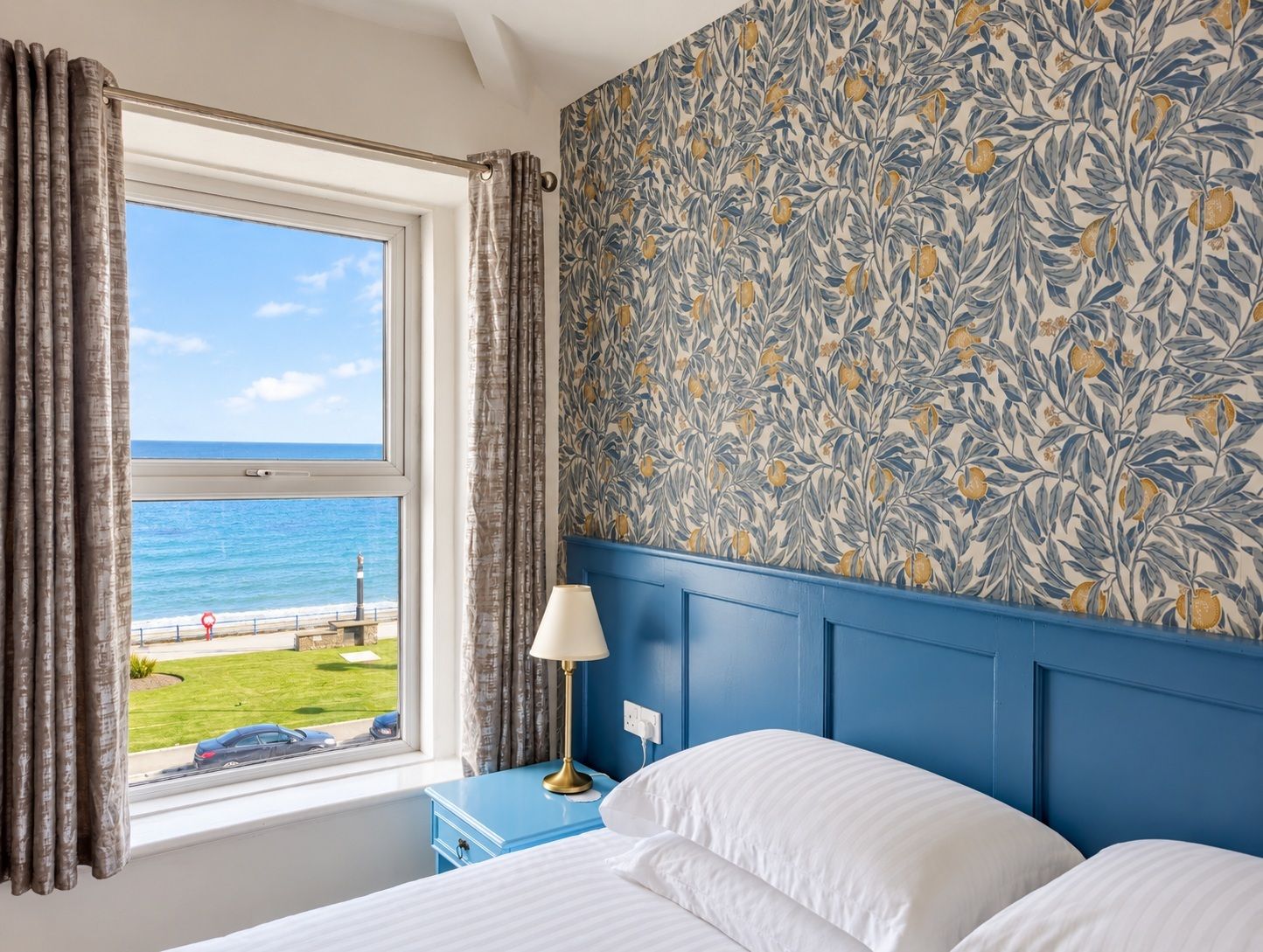 Habitación de hotel con cama, mesita de noche con lámpara, ventana con cortinas y pared con estampado floral.
