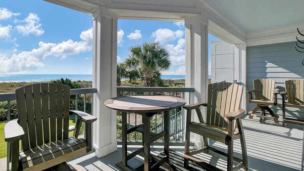 Un espacio de estar al aire libre techado con sillas Adirondack y una mesa, con vistas a una playa y palmeras.