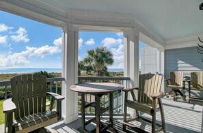 Un espace extérieur aménagé, abrité et doté de chaises Adirondack et d’une table, offrant une vue sur une plage et des palmiers.