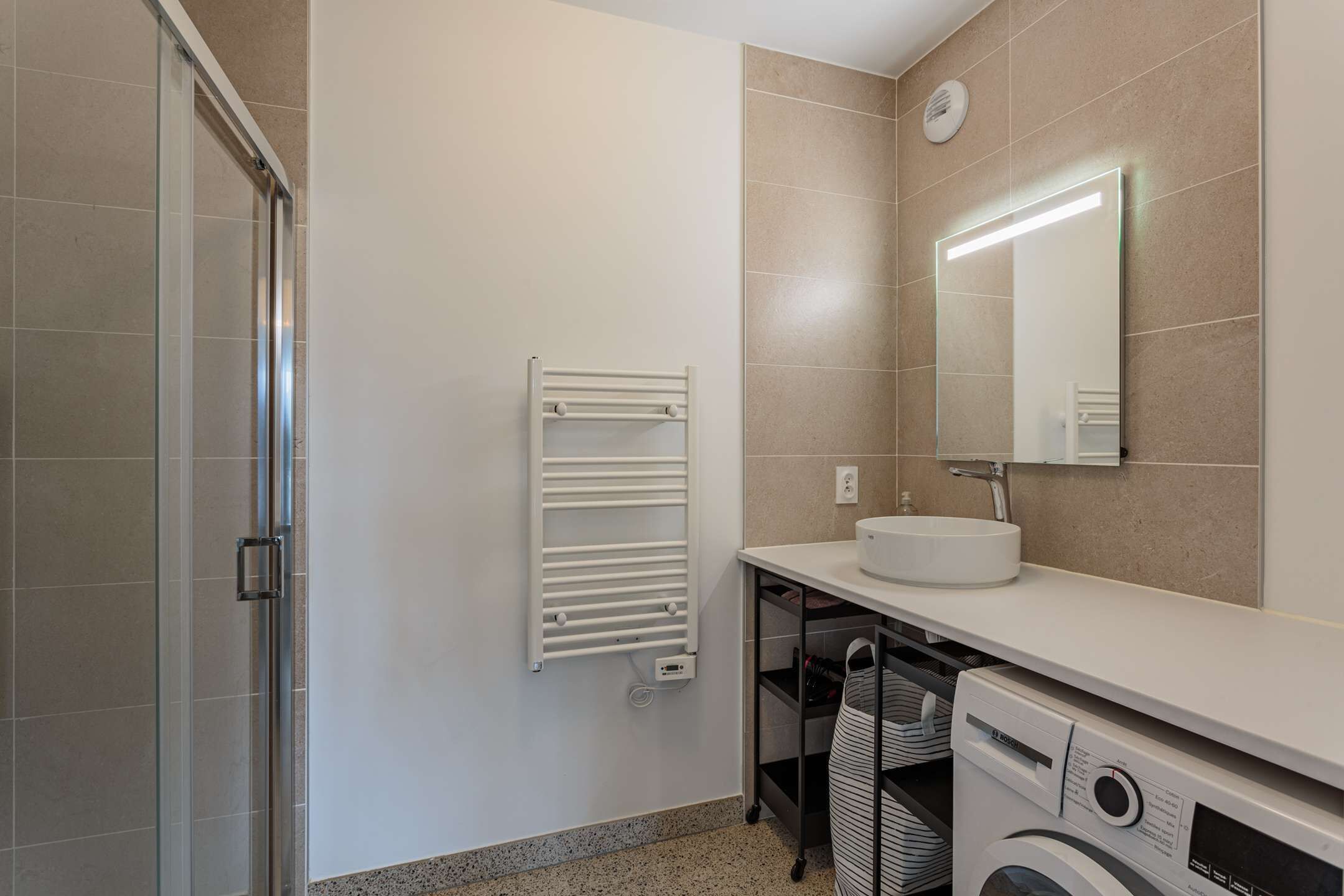 A modern bathroom with a glass shower enclosure, a white towel rack, a sink with a mirror, and a built-in washing machine.