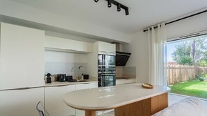 Una cocina moderna con una isla central, una zona de comedor con una silla transparente y vistas a un patio trasero a través de amplios ventanales.