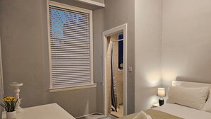 Ett hotellrum med en säng, ett sängbord, en lampa och ett fönster med persienner.