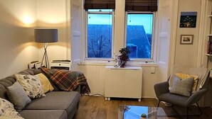 Ett vardagsrum med en soffa, en fåtölj och ett soffbord i glas. Det finns ett fönster med persienner, en lampa och ett element.