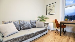 A living room with a worn sofa, a dining table with chairs, a potted plant, and a framed artwork on the wall.
