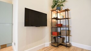 A room with a flat-screen TV mounted on the wall, a wooden floor, and a metal shelving unit with various items including a plant.