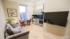 A compact living space with a sofa, dining table, chairs, a washing machine, kitchen appliances, and a flat-screen TV.