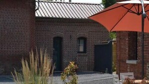 Ein Gebäude aus Backsteinen mit einer Metallbedachung, Außenbestuhlung mit einem roten Sonnenschirm und Pflanzkübeln.