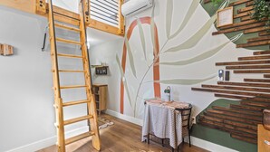 Una habitación con una escalera de madera, un aire acondicionado instalado en la pared, una mesa con mantel a rayas y una pared con diseño de hojas.