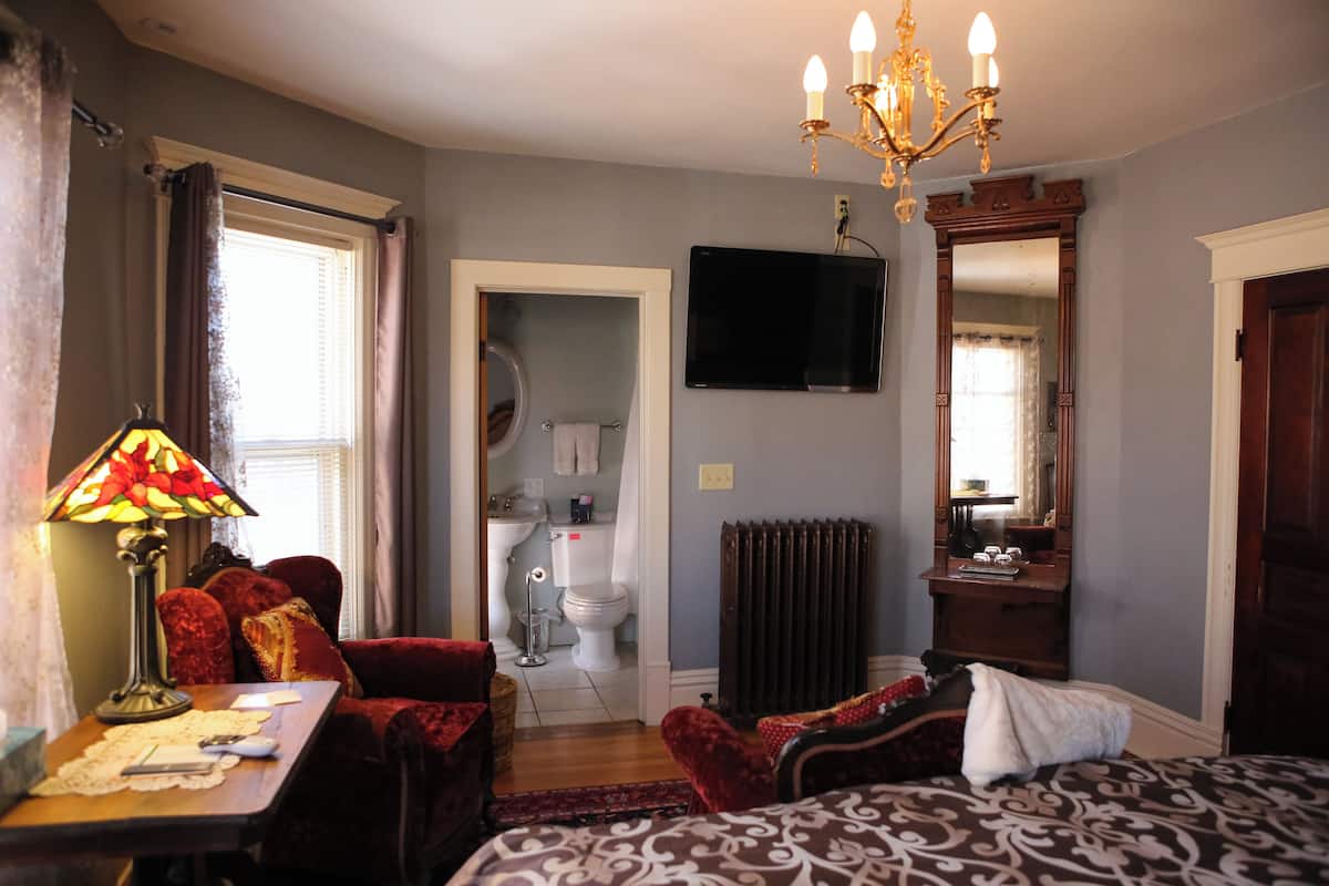 A bedroom with a bed, a TV, a chandelier, a desk with a lamp, and a bathroom visible through an open door.