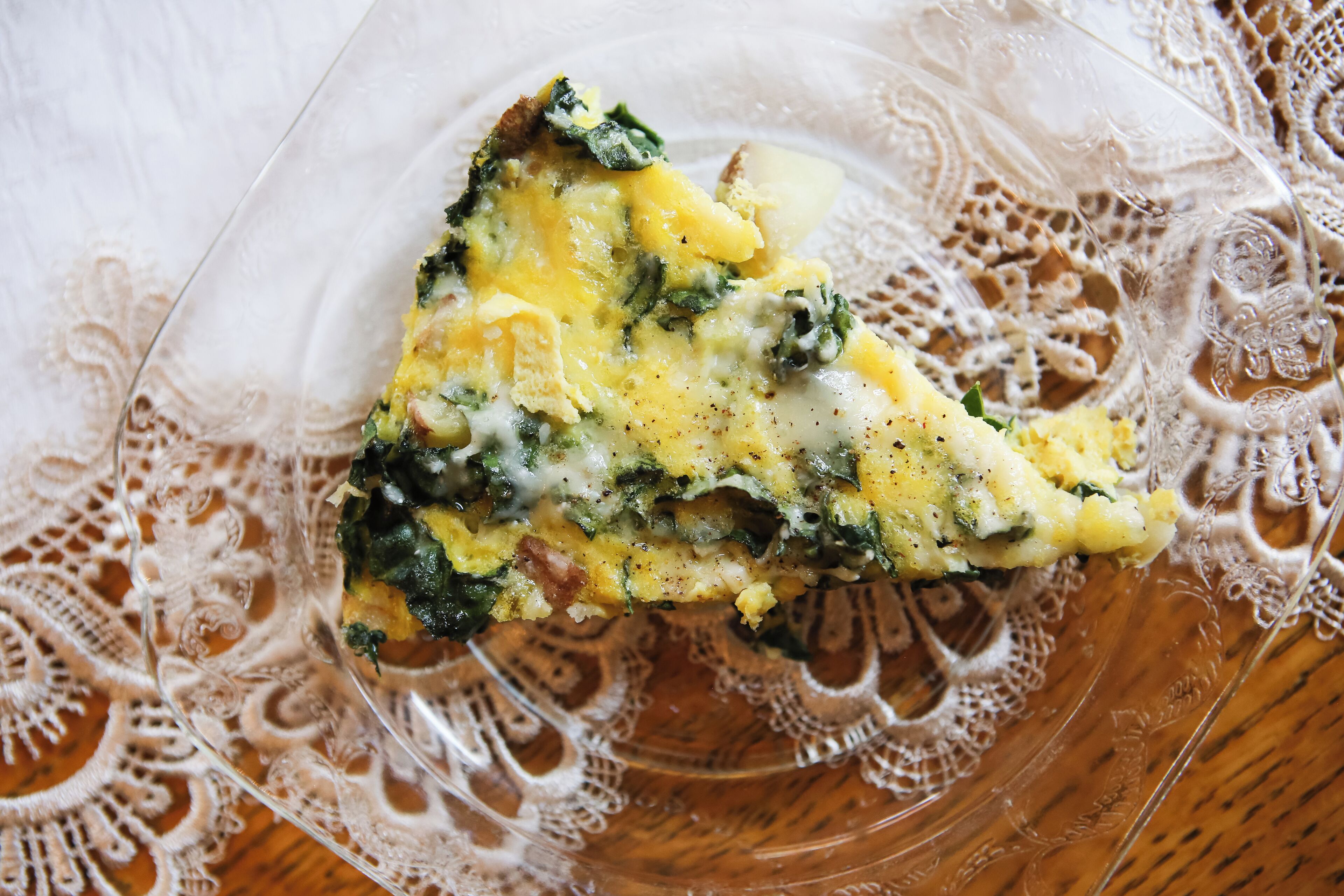 A slice of omelette on a clear glass plate with a lace tablecloth underneath.
