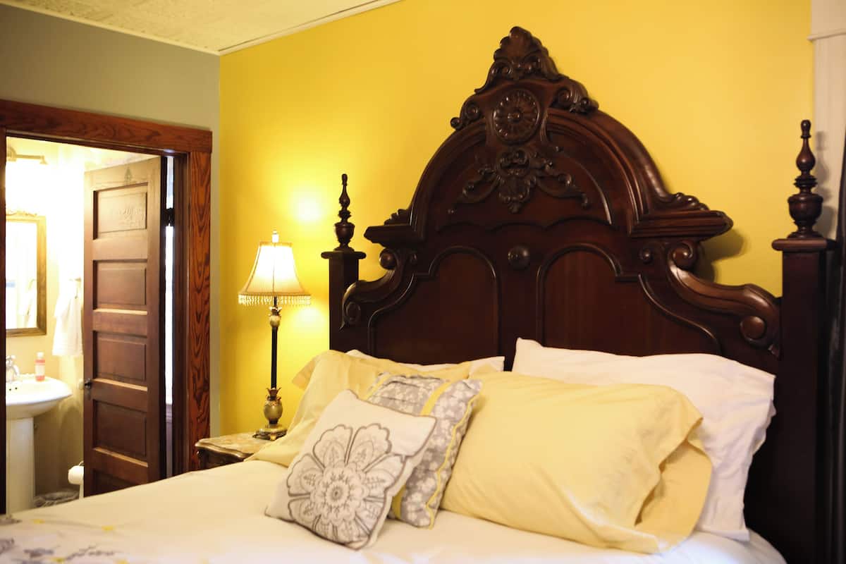 A bedroom with a wooden headboard, a bedside table with a lamp, and a bed with yellow bedding and decorative pillows.