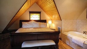A bedroom with a wooden ceiling, a large bed, a bench, a nightstand, a lamp, and a bathtub. - B&B on Broadway (Decorah)