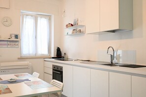 Une cuisine de taille réduite avec des meubles blancs, un four encastré, un évier et une fenêtre avec des rideaux.