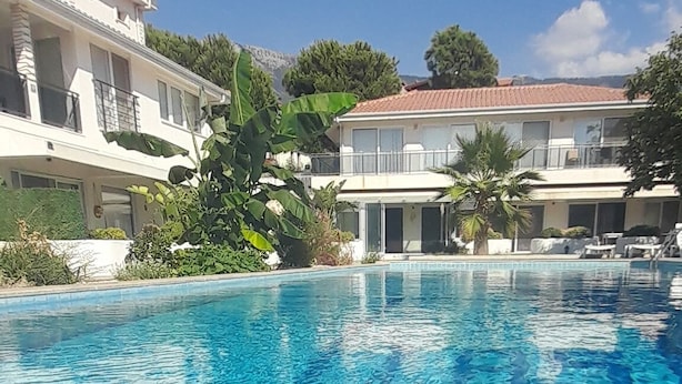En klar swimmingpool med flisekant, omgivet af frodig vegetation og en bygning med flere etager og altaner.