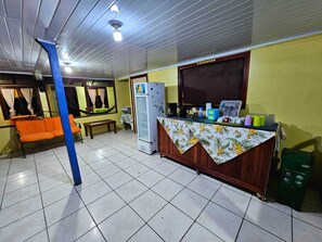 Une petite pièce fermée, dotée d’un sol carrelé, d’un canapé, d’une table, d’un réfrigérateur et d’un comptoir sur lequel sont disposés divers objets.