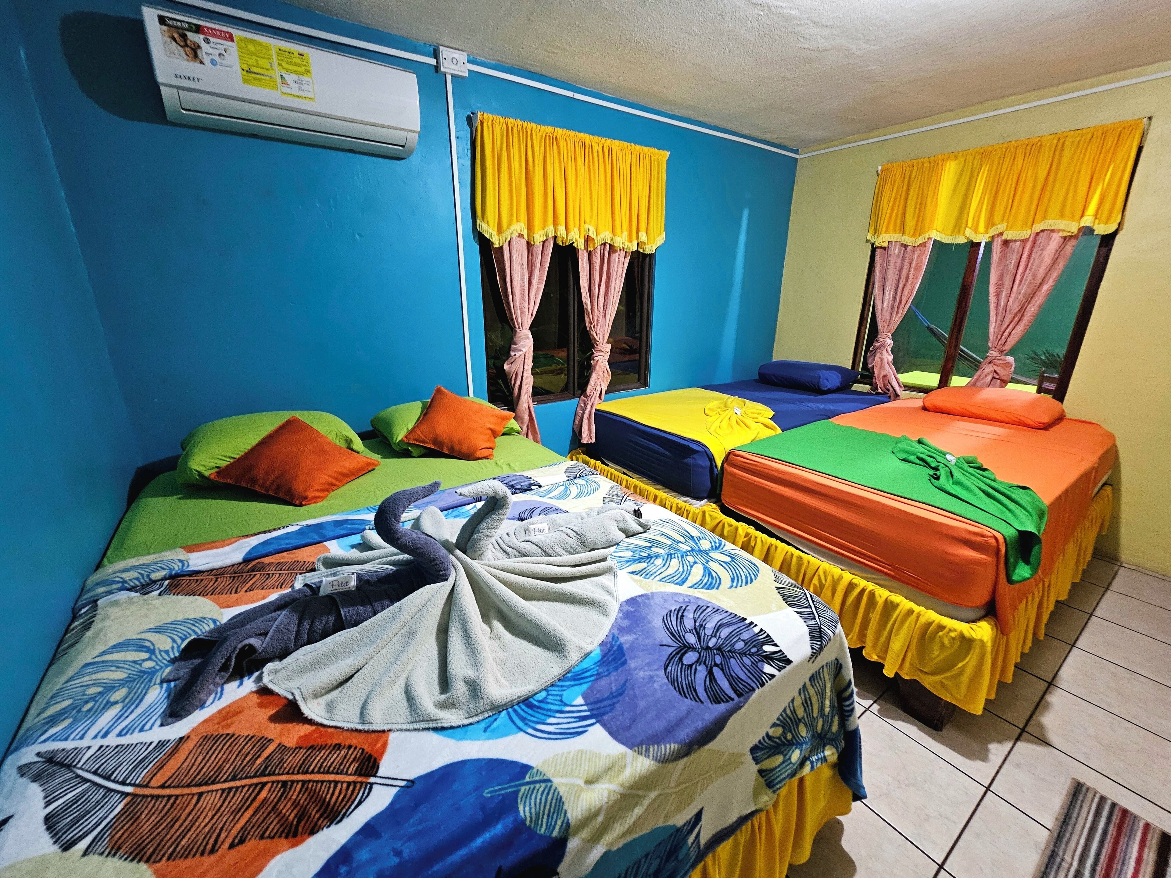 Habitación con dos camas, cada una con ropa de cama y almohadas coloridas. Se ve una ventana con cortinas amarillas y un aire acondicionado de pared.