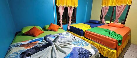 Habitación con dos camas, cada una con ropa de cama y almohadas coloridas. Se ve una ventana con cortinas amarillas y un aire acondicionado de pared.
