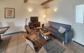 Ein Wohnzimmer mit einer Couch, zwei Korbstühlen, einem Couchtisch, einer Gitarre, einem Fernseher und einem gemusterten Teppich.