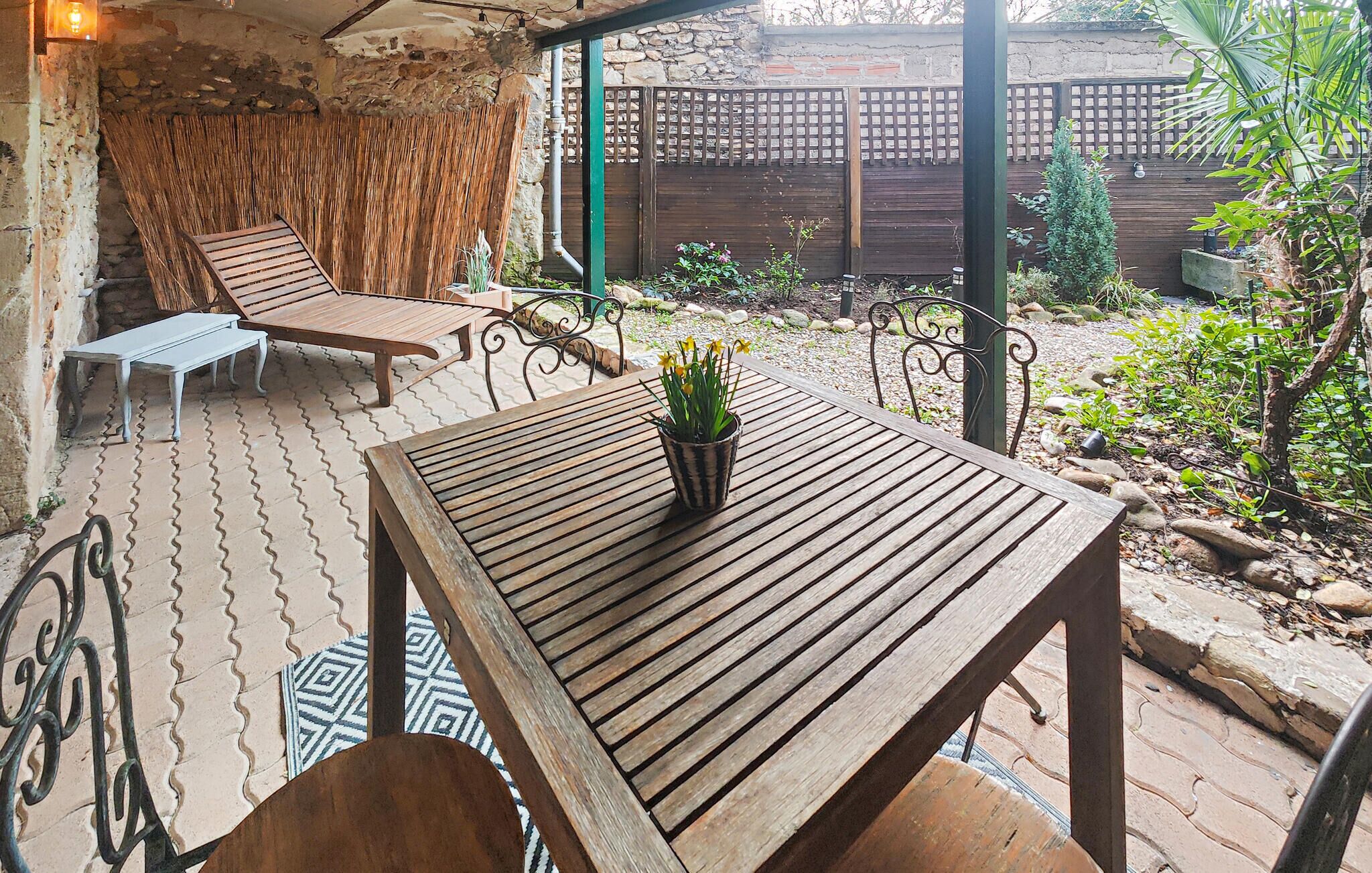 A wooden table with a potted plant, a metal chair, and a lounge chair in a covered outdoor area with a stone wall and a wooden fence.