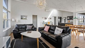 Un salón moderno con un sofá de cuero negro, una mesa de centro blanca y una araña. Se ve un área de cocina con una mesa de comedor y sillas al fondo.