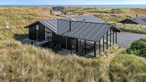 Una casa moderna con un porche de cristal, rodeada de pastos altos y dunas, con vistas al paisaje costero.