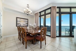 A dining room with a large table and chairs, a chandelier, a framed picture on the wall, and a view through large windows.