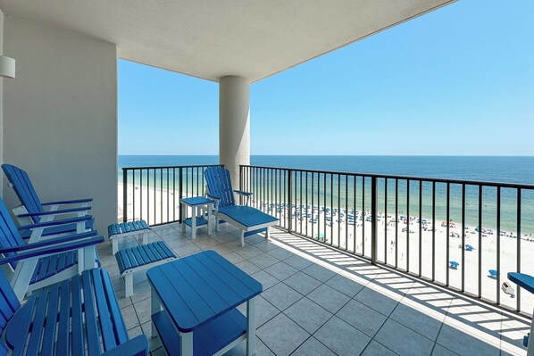 Un balcon avec des chaises et des tables bleues, donnant sur une plage parsemée de parasols et où se trouvent des gens.