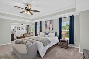 A bedroom with a large bed, a ceiling fan, a nightstand, and a window with curtains.