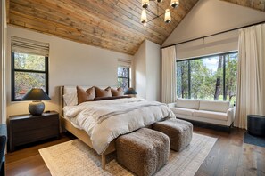 Quarto com teto de madeira, uma cama grande com cabeceira, mesa de cabeceira, luminária, janela com vista para árvores e um sofá junto a uma janela ampla.