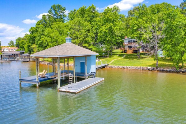 Una casa flotante amarrada con cubierta techada y un pequeño porche, rodeada por un lago tranquilo y abundante vegetación.