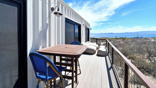 A deck with a table and chairs, overlooking a landscape with mountains and a body of water. - Funnest house in Baja! Game nights w/ smart darts, shuffleboard, & video games. Enjoy a new 8 person hot tub, lounge on multiple decks w/ firepits, fast Wifi ( 250 mb+)w/ Starlink backup. Use of 2 SUPs.  Daily housekeeping optional. (El Sargento)