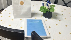 A round table with a white tablecloth featuring gold polka dots, a blue book titled "House Manual," a white paper bag with a gold logo, and a small potted plant with blue and white flowers. - Charming old 3-bedroom with WiFi, & central heat & air in Cabot. NEWLY Renovated (Cabot)