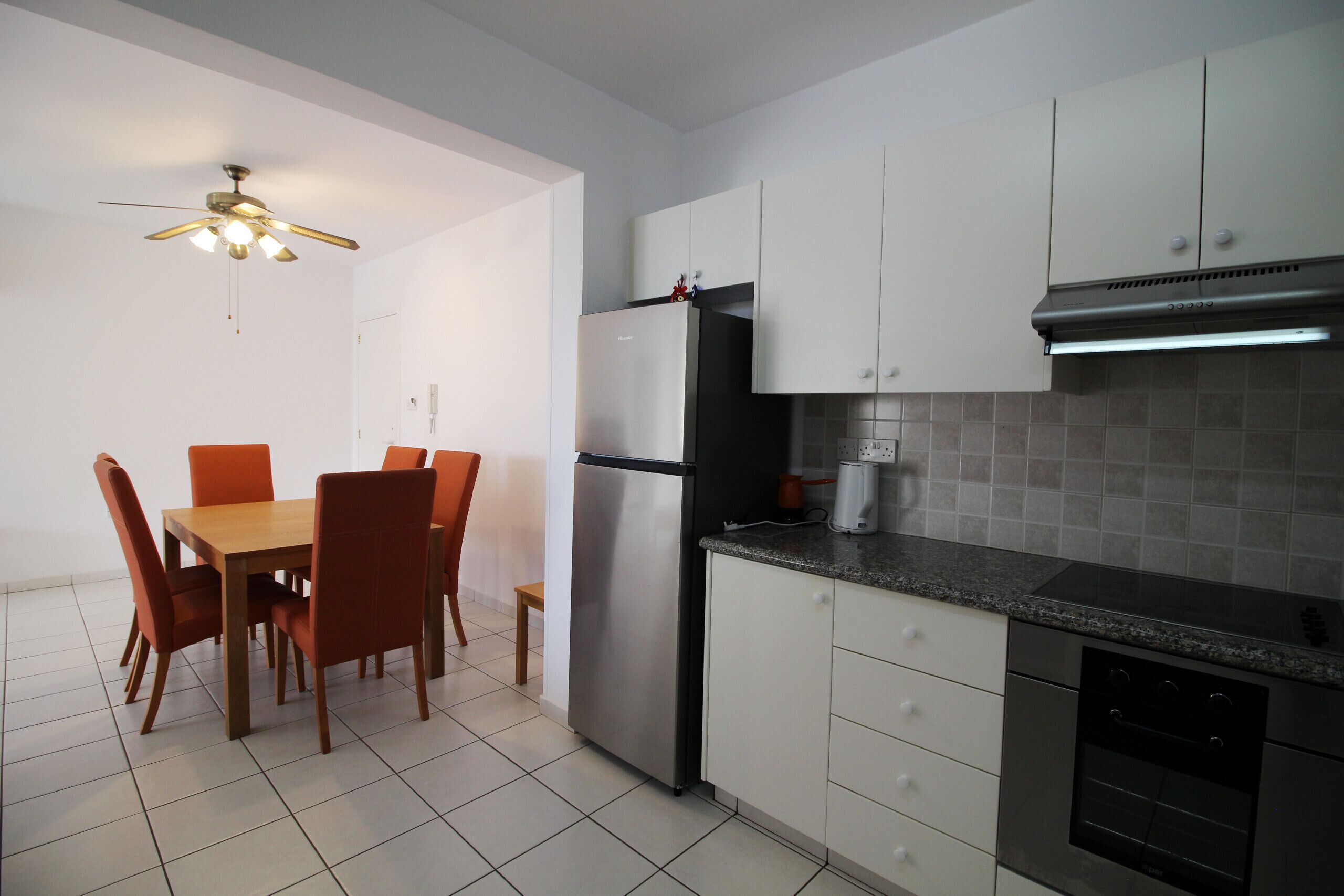 Une cuisine moderne avec des meubles blancs, un réfrigérateur en acier inoxydable, un four noir et un coin repas comprenant une table en bois et des chaises orange.