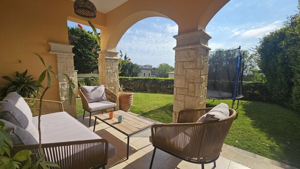 Un patio al aire libre con mobiliario de mimbre, una mesa y vistas a un césped cuidado y edificios a la distancia.