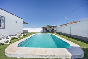 En rektangulär swimmingpool med en vit poolkant, omgiven av en gräsyta och en vit byggnad med en balkong.