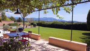 An outdoor patio with a table and chairs, surrounded by greenery and a view of a well-maintained lawn and distant mountains. - In Violès, Vaucluse, exceptional vacation home, air-conditioned, heated pool. (VIOLÈS)