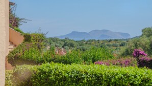 A lush garden with a variety of plants and flowers, a building with a beige facade, and mountains in the background. - Apartment 'Avocado' with Shared Pool, Private Garden & Air Conditioning (Maltineddu)