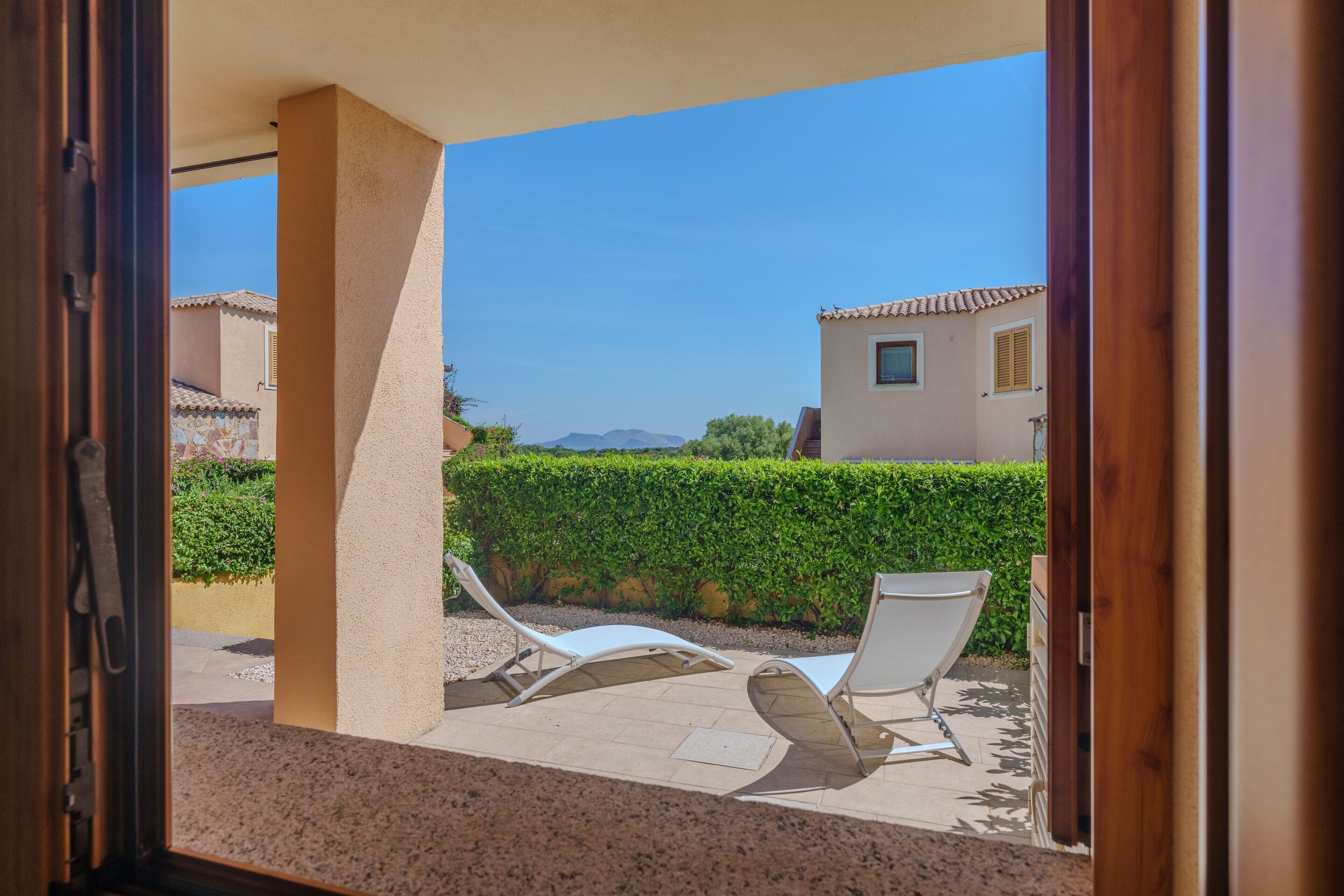 A view through an open door to a balcony with two lounge chairs, a beige wall, and a hedge in the background.