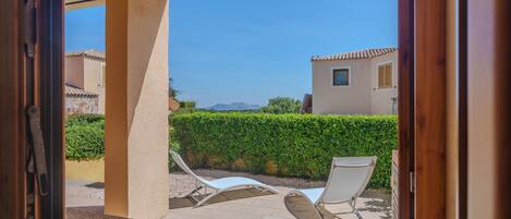 A view through an open door to a balcony with two lounge chairs, a beige wall, and a hedge in the background.