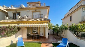 A residential building with a patio area featuring a tiled walkway, green lawn, and blue lounge chairs. - Holiday Home 'Relax Inn' with Sea View, Shared Pool & Wi-Fi (Pilar de la Horadada)