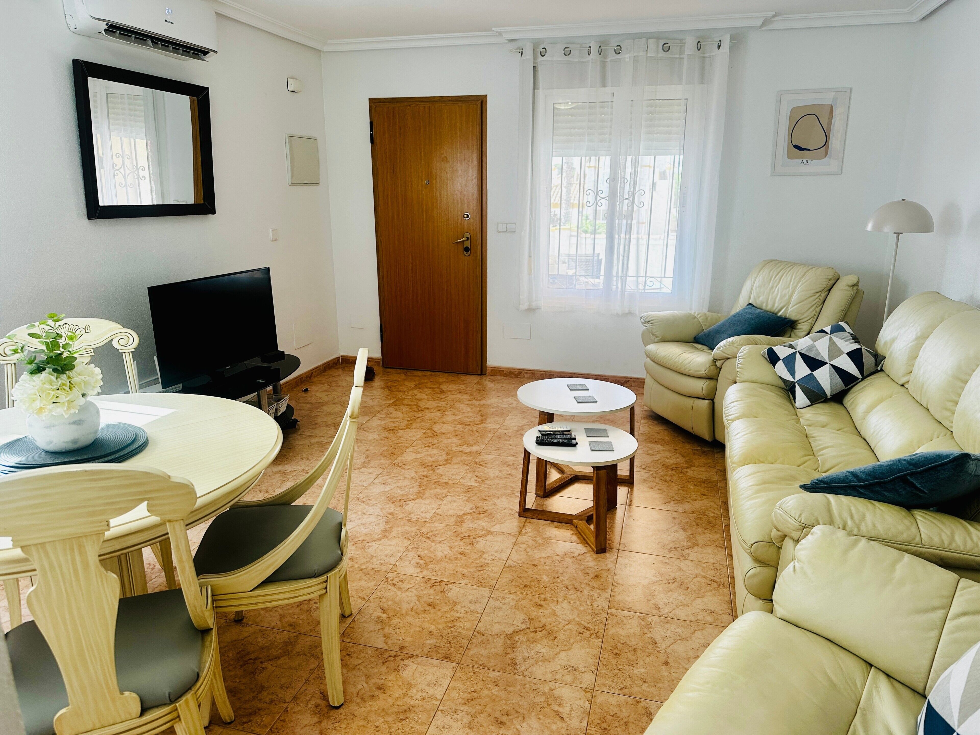 Een woonkamer met een beige bank, een eettafel met stoelen, een televisie en een deur.