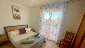 A hotel room with a bed, a chair, a nightstand, a window with curtains, and a painting on the wall. - Holiday Home 'Relax Inn' with Sea View, Shared Pool & Wi-Fi (Pilar de la Horadada)