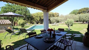 An outdoor patio with a table set for dining, surrounded by chairs, a pool, and a covered area with a wooden roof. - Stone landhouse in scenic garden, heated pool, in L'Isle-sur-la-Sorgue. (L'ISLE-SUR-LA-SORGUE)
